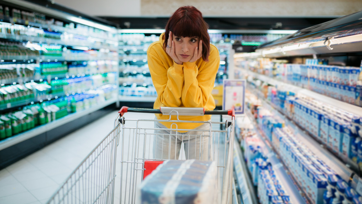 Young woman with red hair buying groceries in a local supermarket, feeling depressed by the high prices in the store and leaning on her shopping cart