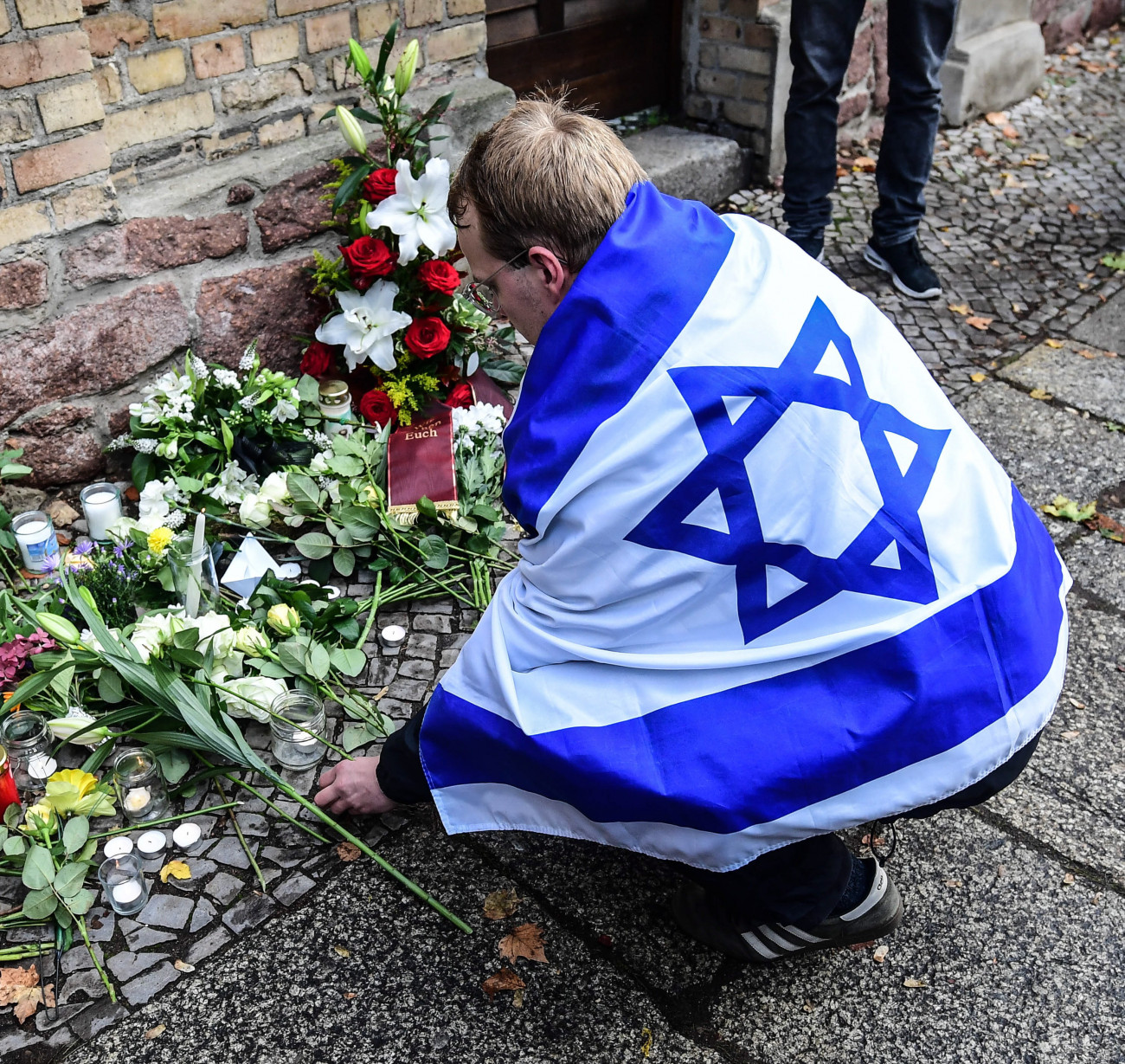 Halle, 2019. október 10.
Virágot helyez el egy izraeli zászlóba burkolózott férfi a hallei zsinagógánál a németországi városban előző nap elkövetett lövöldözés utáni napon, 2019. október 10-én. A tettes, egy 27 éves német neonáci férfi a sisakjára szerelt kamerával élőben közvetítette a zsinagóga elleni sikertelen támadását, majd azt, amint az utcán hátulról lelő egy fiatal nőt, aki éppen arra járt, majd autójával egy közeli török büféhez hajtott, amelyben lelőtt egy férfit, és elmenekült. Hallétól nagyjából 30 kilométerre fogták el, miután nekihajtott egy teherautónak. MTI/EPA/Filip Singer
MTI/EPA/Filip Singer