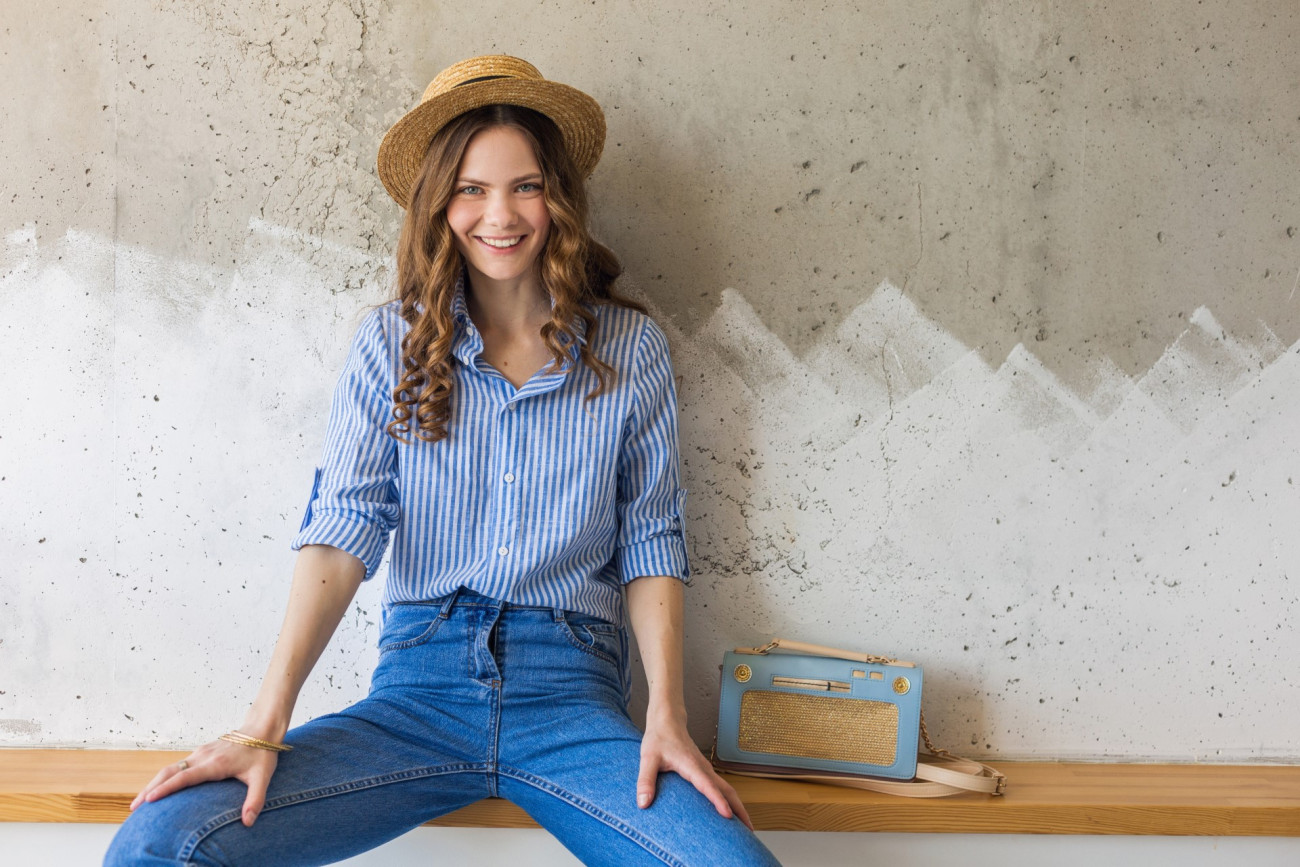 young attractive stylish woman sitting at wall background, straw hat, wearing blue cotton shirt, jeans, casual outfit, hipster style, spring summer fashion trend, smiling face, happy mood, relaxed