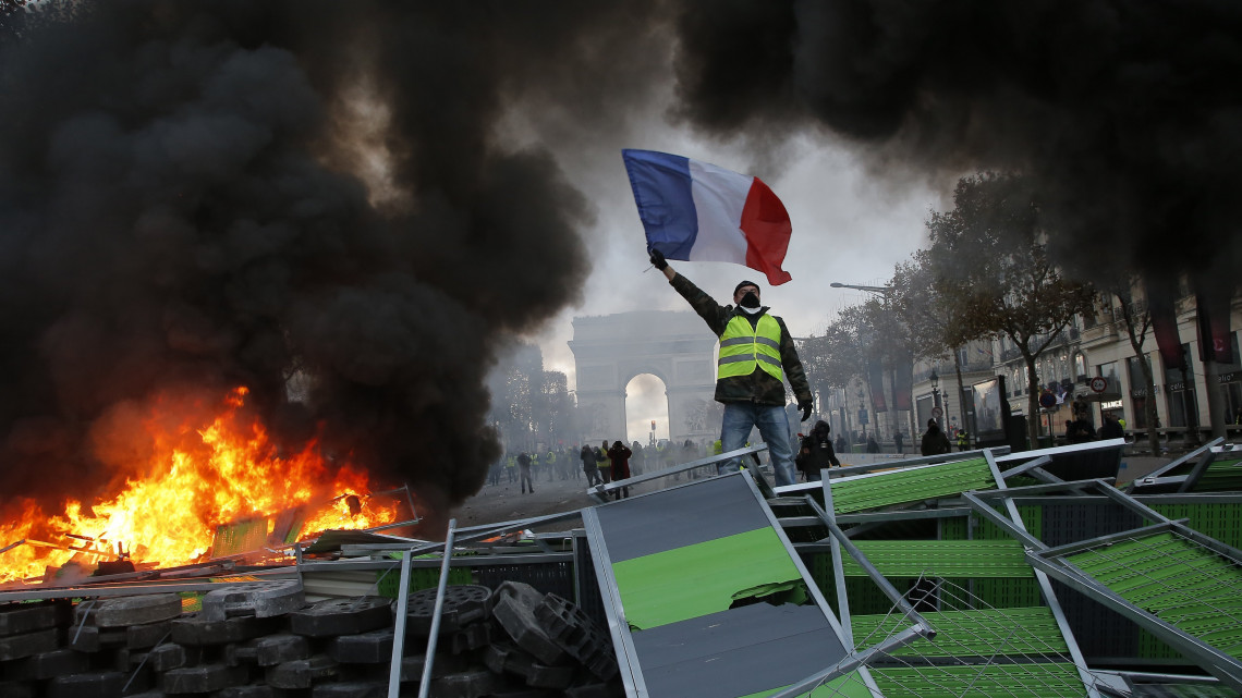 A francia lobogót lengeti egy láthatósági mellényt viselő tiltakozó égő úttorlasznál az üzemanyagadó tervezett emelését ellenző tüntetésen a Champs-Elysées sugárúton Párizsban 2018. november 24-én. Országszerte csaknem 81 ezren vonultak utcákra békés tüntetéseken, a fővárosban 8 ezren tiltakoztak az erőszakossá fajult megmozduláson.