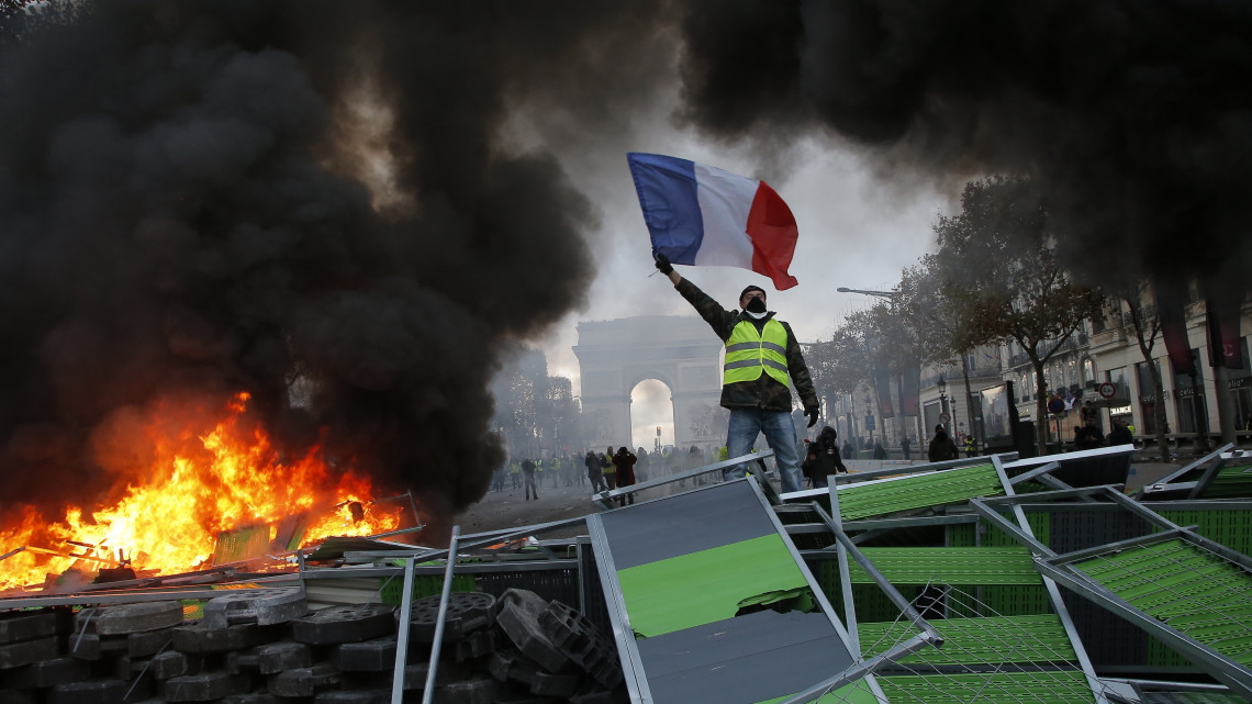 A francia lobogót lengeti egy láthatósági mellényt viselő tiltakozó égő úttorlasznál az üzemanyagadó tervezett emelését ellenző tüntetésen a Champs-Elysées sugárúton Párizsban 2018. november 24-én. Országszerte csaknem 81 ezren vonultak utcákra békés tüntetéseken, a fővárosban 8 ezren tiltakoztak az erőszakossá fajult megmozduláson.
