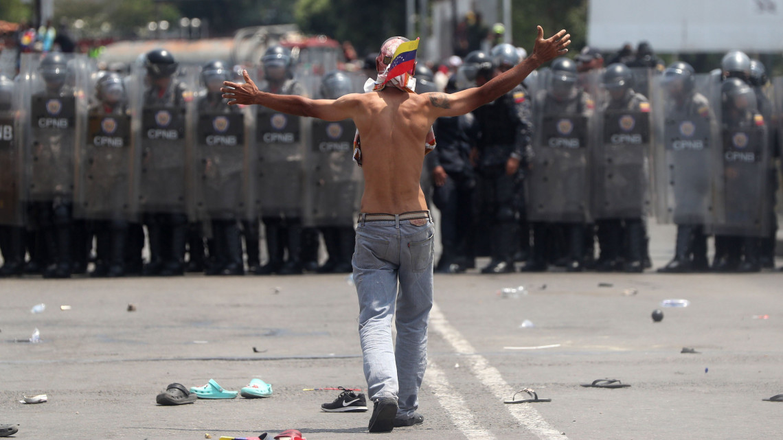 Félmeztelen tüntető a Simón Bolívar hidat lezáró venezuelai rohamrendőrök előtt a határ kolumbiai oldalán lévő Cúcutában 2019. február 23-án. Nicolás Maduro venezuelai elnök előző nap rendelte el a Kolumbiával közös határ egyes átkelőinek lezárását, hogy megakadályozza az Egyesült Államokból érkező segélyszállítmány bejuttatását az országba. Venezuelában hatalmi harc zajlik Maduro és a magát ideiglenes elnökké nyilvánító Juan Guaidó parlamenti elnök között. Guaidó, aki január 23-án kikiáltotta magát Venezuela ideiglenes elnökének, az Egyesült Államok és több más nyugati ország támogatását élvezi.