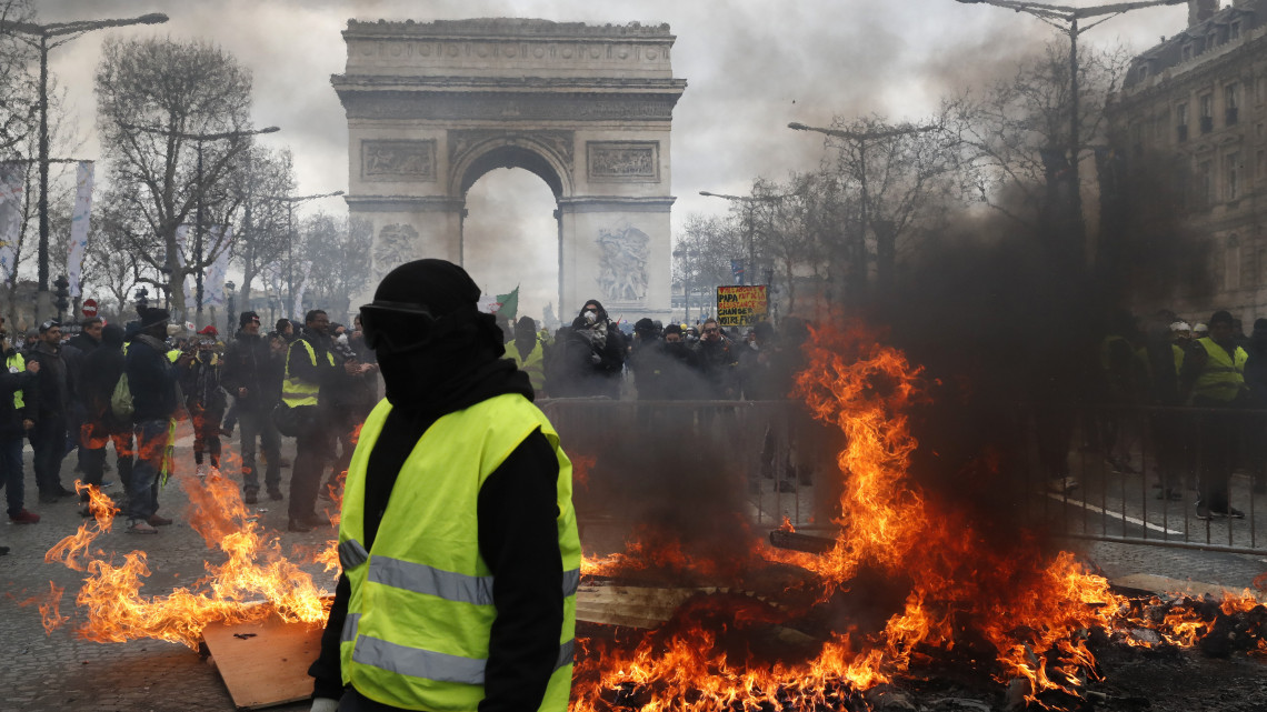Égő úttorlasznál tüntetnek a francia kormány szociális és adópolitikája ellen tiltakozó sárgamellényesek a párizsi Diadalívnél 2019. március 16-án, a tizennyolcadik egymást követő hétvégi tüntetésen. Az üzemanyagadó emelése ellen 2018. november 17-én kezdődött sárgamellényes mozgalom azóta több szociális jellegű követelést fogalmazott meg és Emmanuel Macron államfő távozását követeli.