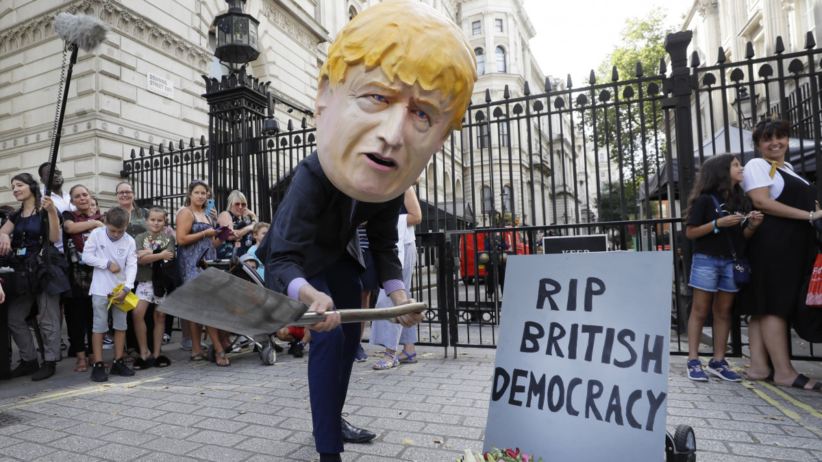Boris Johnson brit miniszterelnököt ábrázoló maszkban jelképesen ássa a sírját egy tiltakozó egy Nyugodj békében, brit demokrácia! feliratú sírkő előtt a londoni kormányfői rezidencia, a Downing Street 10. közelében 2019. augusztus 28-án.