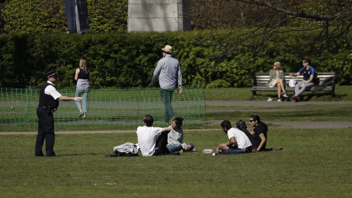 A gyepen ülő társaságot szólít fel mozgásra egy rendőr a londoni Greenwich Parkban, a háttérben az üzleti negyed, a Canary Wharf felhőkarcolói 2020. április 5-én. A koronavírus-járvány miatt a brit kormány kijárási korlátozást vezetett be, amelynek értelmében napi egy testedzés is engedélyezett, de az egyhelyben ülés, napozás a közterületeken nem. A rendelet megszegése miatt egy másik londoni parkot bezárattak a hatóságok.