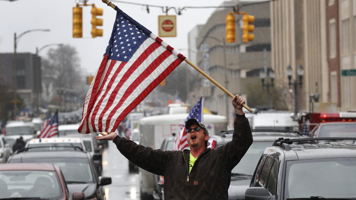 Amerikai zászlót lenget a koronavírus-járvány miatt bevezetett korlátozó intézkedésekkel elégedetlen emberek autói között egy férfi Michigan állam törvényhozásának épülete előtt Lansingban 2020. április 15-én.