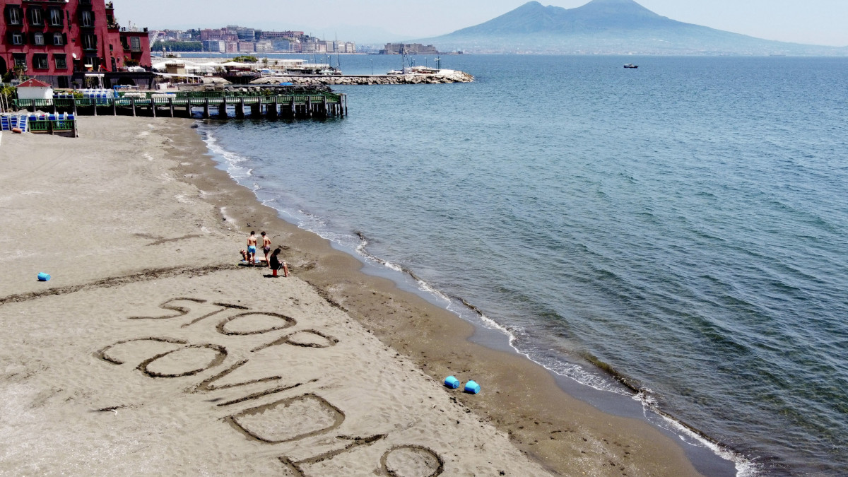 Állítsuk meg a koronavírust! jelentésű feliratot írt a homokba egy család Nápoly Posillipo nevű városrészének tengerpartján 2020. május 9-én. Olaszországban a bevezetett óvintézkedéseket fokozatosan négy fázisban oldják fel, április 27-től négy egymást követő hétfőn újabb és újabb korlátozó intézkedéseken enyhítenek.