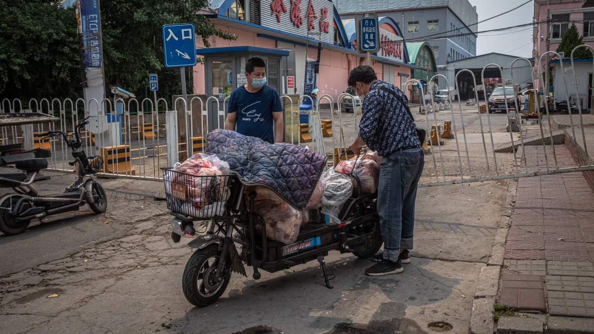 A koronavírus-járvány miatt védőmaszkot viselő emberek egy robogóra rakják fel a csomagjaikat a pekingi Hszinfadi nevű nagybani piac épülete közelében 2020. június 13-án. A piacot lezárták, miután a hatóságok a helyben továbbadott koronavírusos esetek számának megugrását jelentették