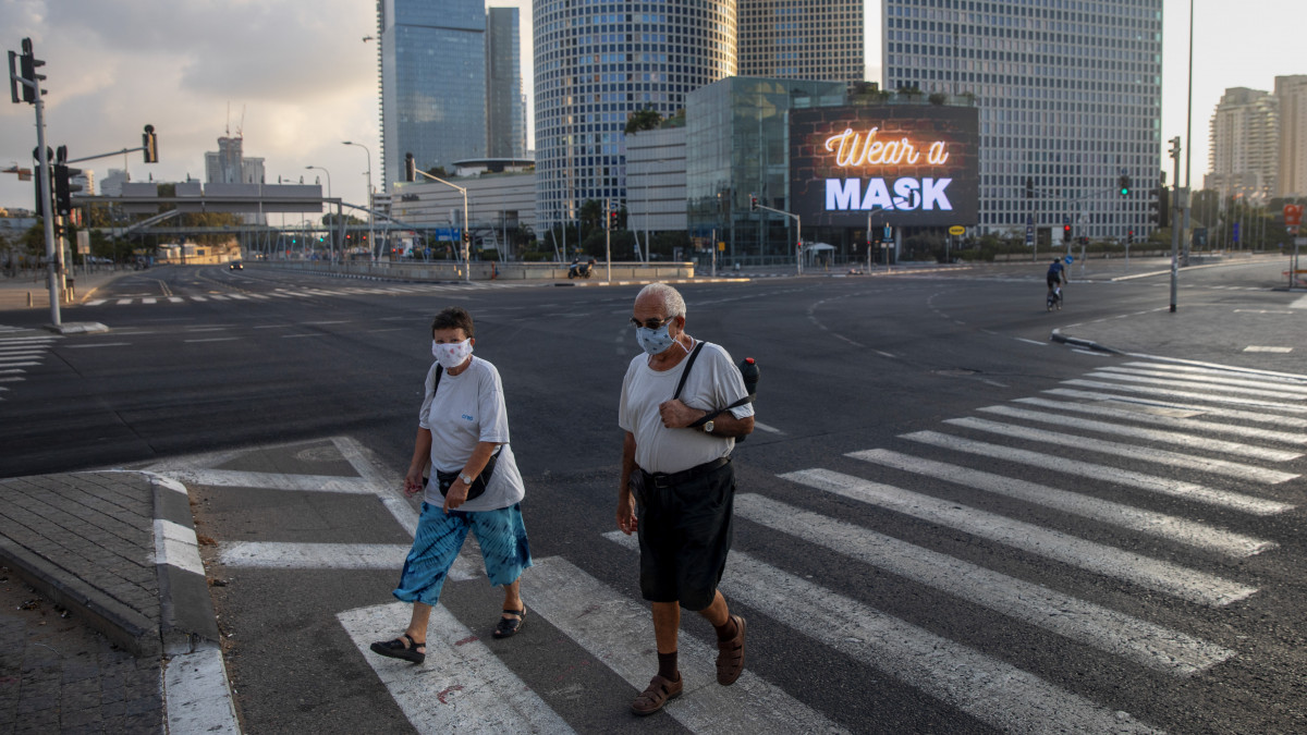 A koronavírus-járvány miatt védőmaszkot viselő emberek mennek át a kijárási korlátozások miatt elnéptelenedett autóúton Tel-Avivban 2020. szeptember 26-án. A háttérben egy világító kijelzőn védőmaszk viselésére figyelmeztető felirat.