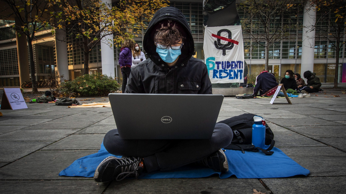 Az online oktatás ellen tiltakozó diákok a földön ülve, a mobileszközeiket használva vesznek részt a tanórákon Milánó belvárosában 2020. november 26-án.