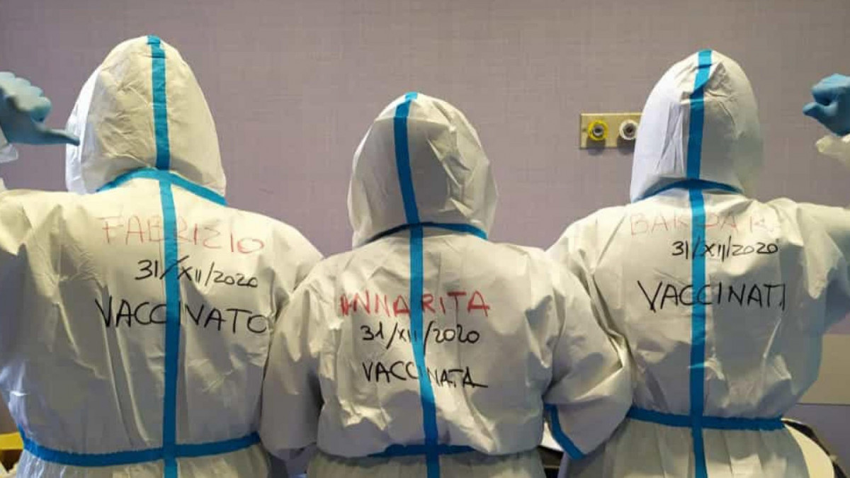 A koronavírus-járvány miatt védőruházatot viselő egészségügyi dolgozók a hátukra írt feliratot mutatják a római Santo Spirito kórházban 2020. december 31-én. A felirat a nevüket, és azt a dátumot mutatja, amikor megkapták a vírus elleni oltást.