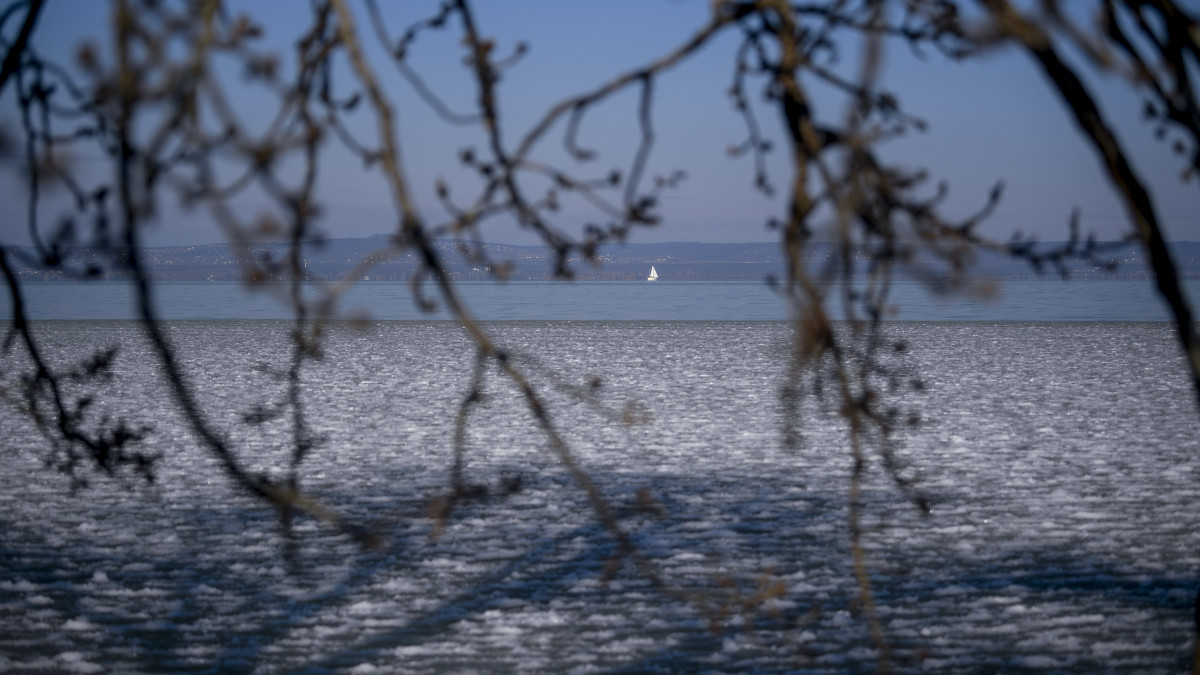 A part mentén jegesedő tó, a háttérben egy vitorláshajó a balatonlellei kikötő felől fényképezve 2021. február 13-án.