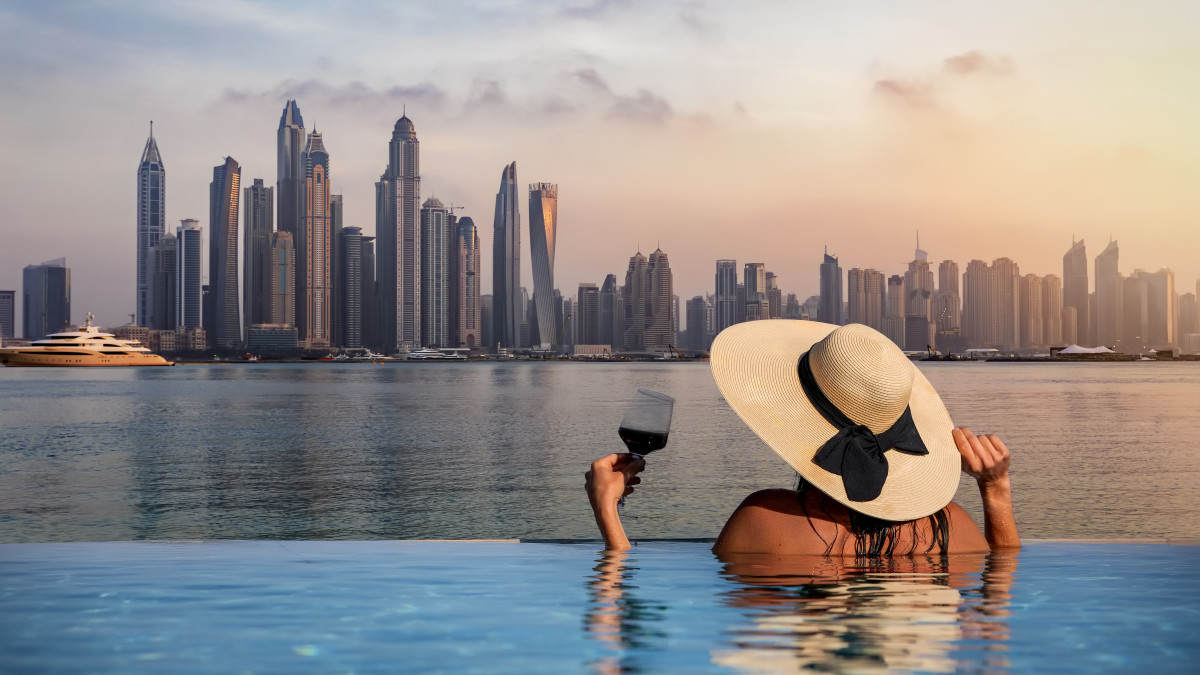 A elegant woman with a hat and a drink in her hand stands at the edge of a infinity pool and enjoys the view to the skyline of the Dubai Marina during sunset, UAE