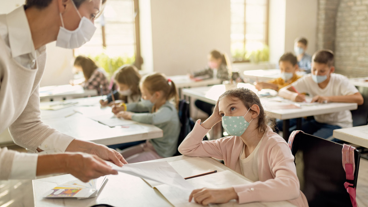 Small girl and her teacher wearing protective face masks while talking about exam papers curing a class at elementary school.