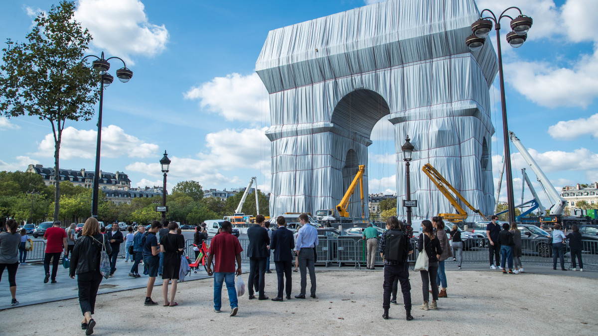 Érdeklődők a párizsi diadalív becsomagolásának utolsó fázisát nézik 2021. szeptember 16-án. Az installációt a néhai Christo bolgár származású csomagolóművész és felesége, alkotótársa, Jeanne-Claude tervei alapján valósították meg 25 ezer négyzetméternyi újrahasznosított ezüstös kék műanyaggal és 3000 méter vörös kötéllel. A hivatalos avatása szeptember 18-án lesz.