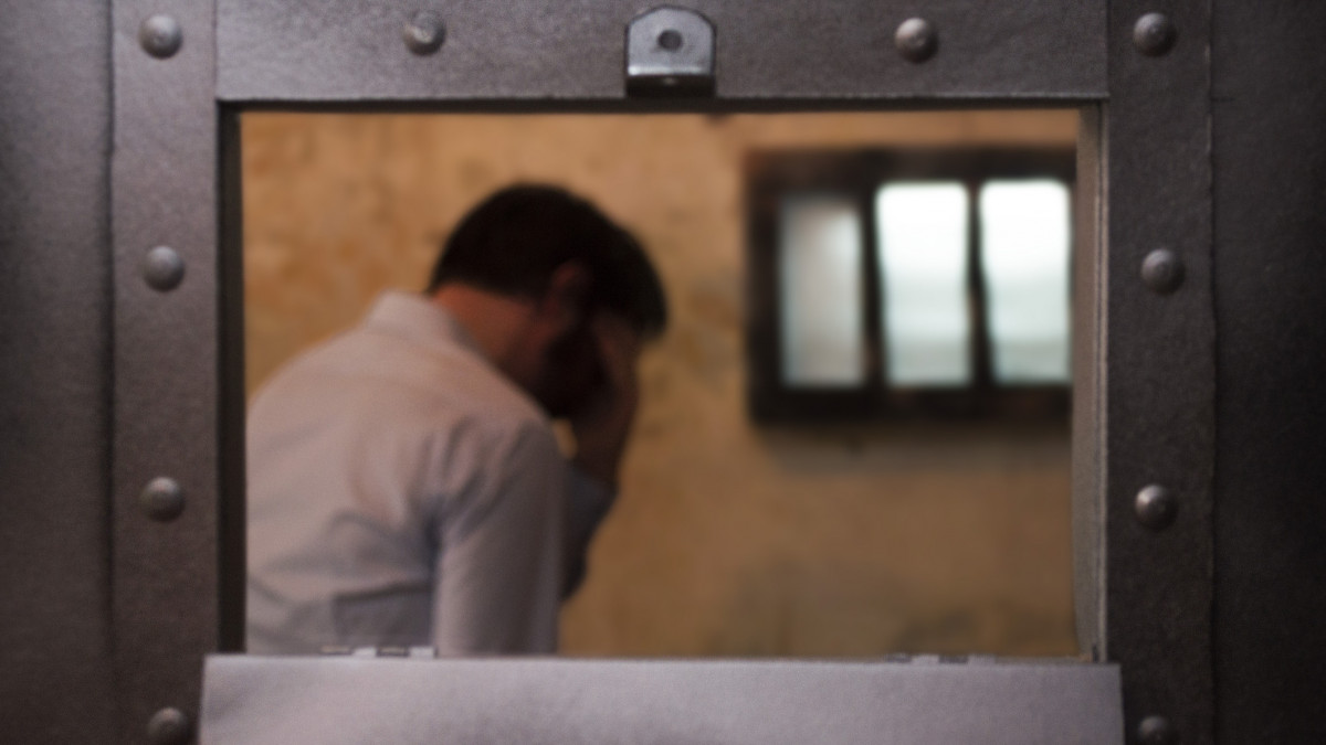 A man stands in a prison cell by a barred window, his head in his hand.