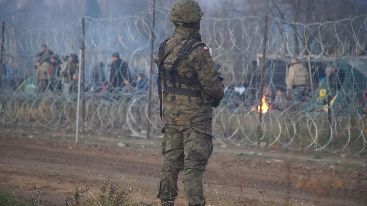A lengyel 16. gépesített hadosztály által közreadott képen az Európai Unióba igyekvő migránsok a lengyel határt védő szögesdrótkerítés fehérorosz oldalán, az előtérben lengyel katona a kelet-lengyelországi Kuznica térségében 2021. november 8-án. A lengyel-fehérorosz határon feszültség alakult ki a Fehéroroszország felől az Európai Unióba igyekvő több ezer migráns miatt. A lengyel hatóságok november 12-i becslése szerint 3-4 ezer migráns tartózkodik a fehérorosz-lengyel határ mentén, és a számuk folyamatosan gyarapodik.