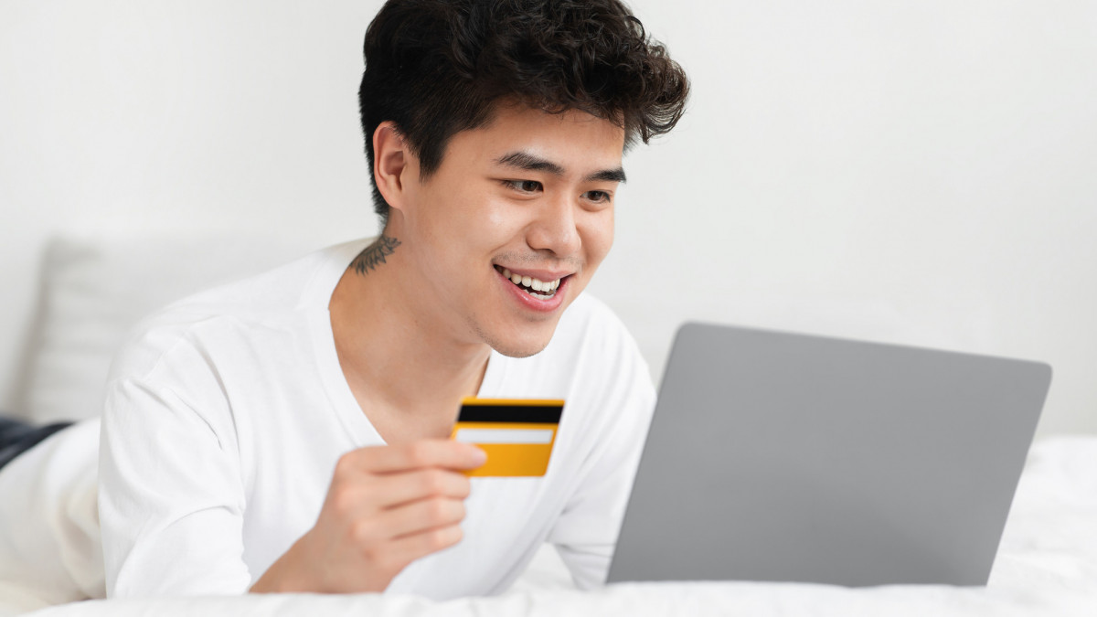Online shopping at home, ordering goods in modern store, covid-19 lockdown. Happy asian young 20s guy in white t-shirt lies on bed in bedroom interior, uses laptop and holds credit card, empty space