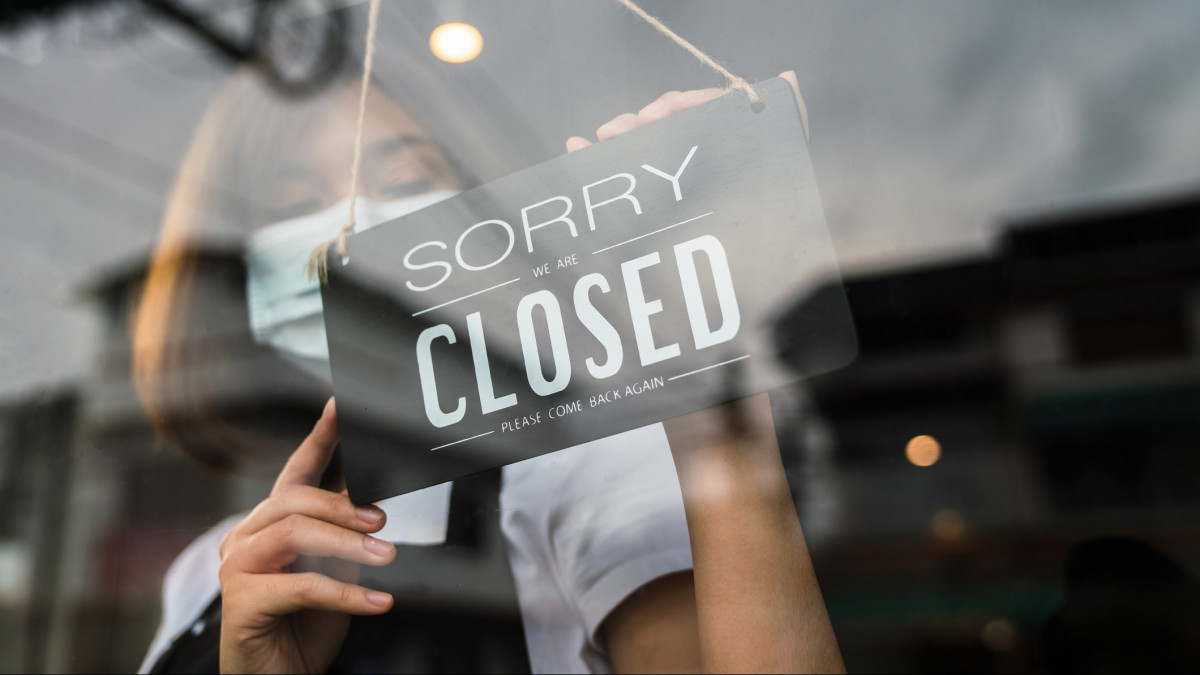 Young asian woman wearing face mask and hand turning closed sign board on glass door in coffee shop and restaurant after coronavirus lockdown quarantine. Business crisis concept.
