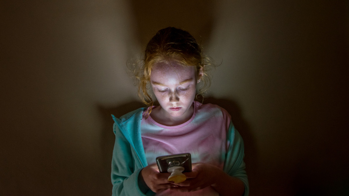 A young long haired redhead girl with face lit up looking into mobile phone screen in a dark room, against a flat wall, with sad facial expression