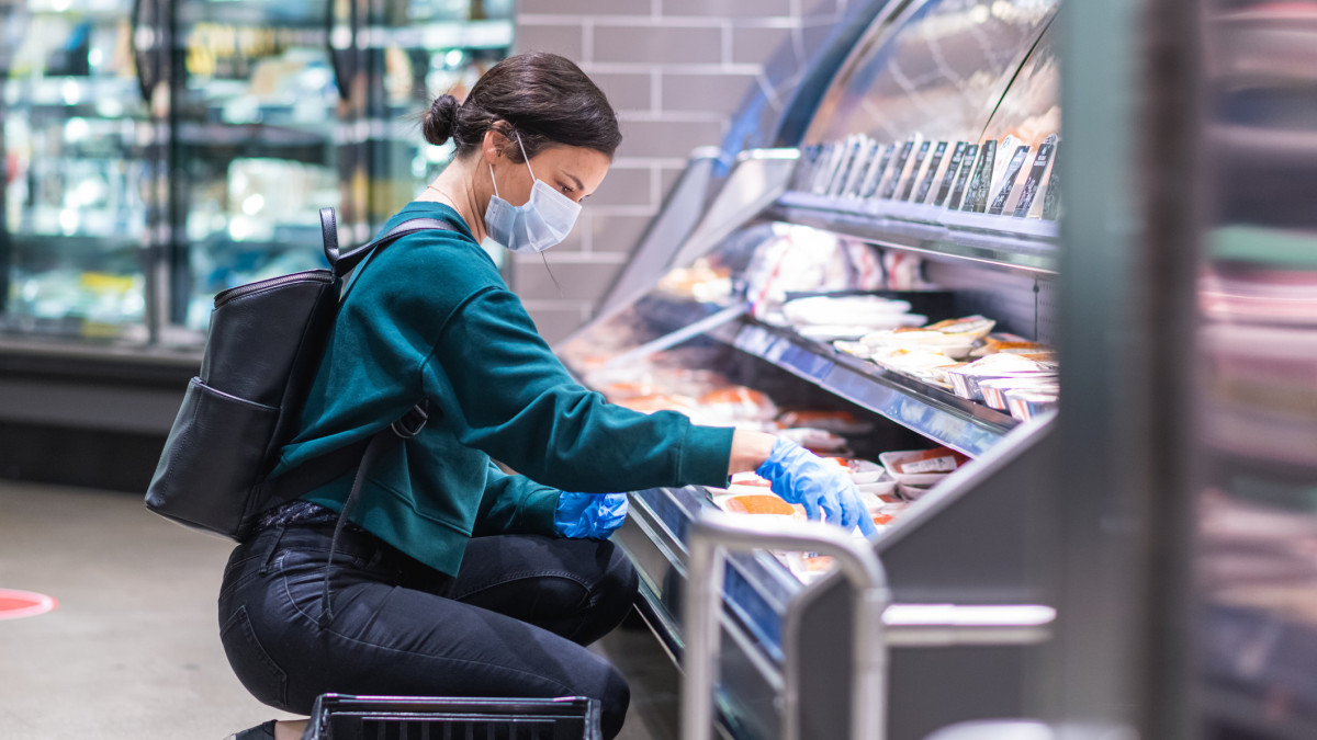 A young woman is shopping in the meat section at a local supermarket during the COVID-19 pandemic. She is wearing a protective face mask and gloves.