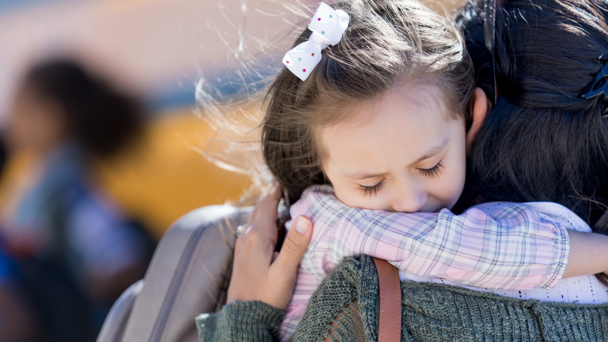 Worried little girl hugs her mom on the first day of school. A school bus is in the background.