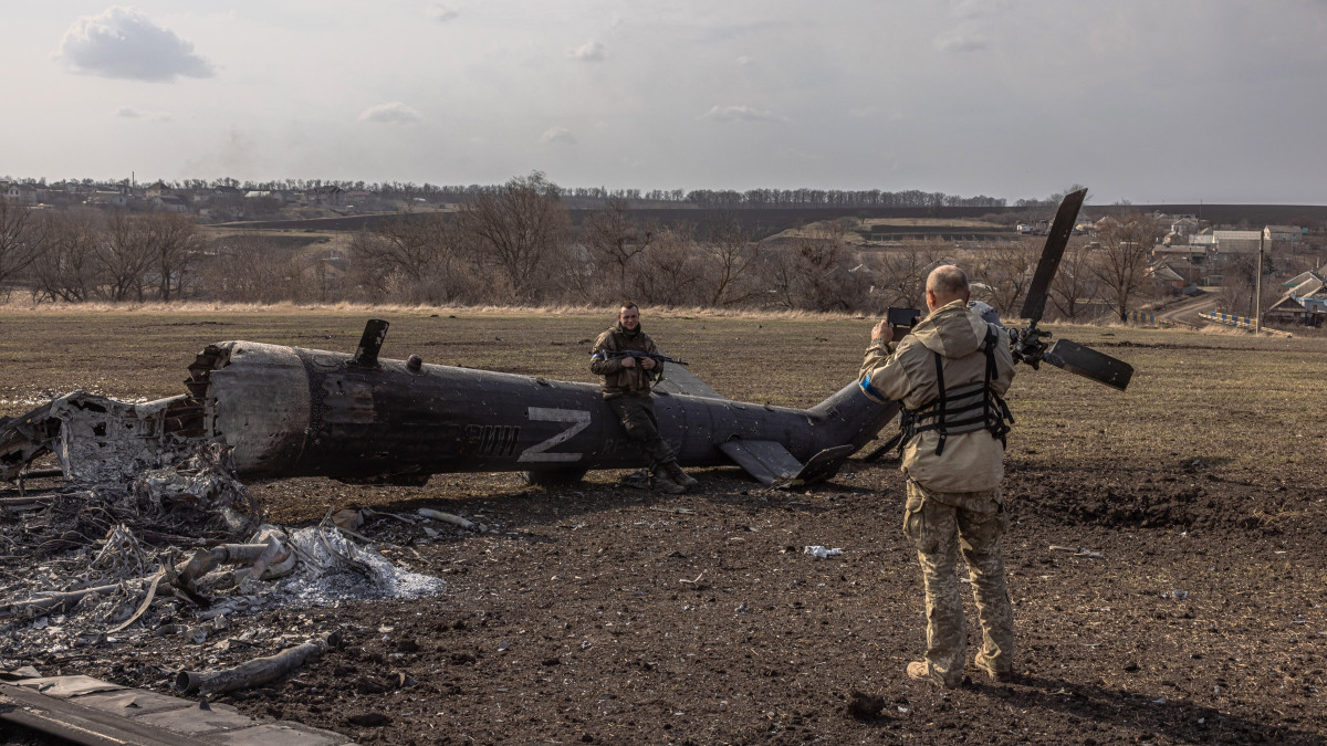 Ukrán katonáról készít képet egyik társa egy megsemmisített orosz helikopter mellett a kelet-ukrajnai Harkiv közelében 2022. március 31-én.