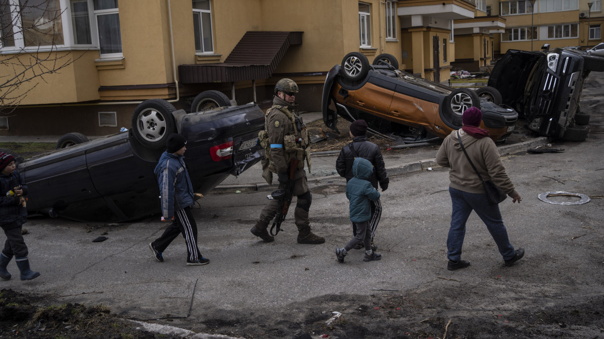 Ukrán katona gyerekeket kísér felborult autók mellett Bucsában, Kijev közelében 2022. április 4-én. Az ukrán hadseregnek mintegy 30 települést sikerült visszafoglalni a főváros körül.