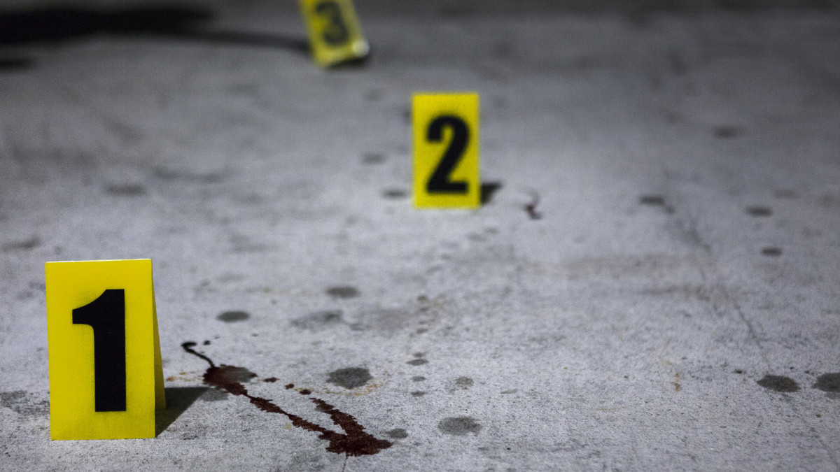Close up of an evidence marker and blood splatter at a crime scene, hand placing more markers in the background.