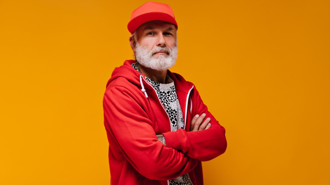 Portrait of man in red cap and hoodie on orange background. Modern guy with grey beard in bright clothes looking into camera..