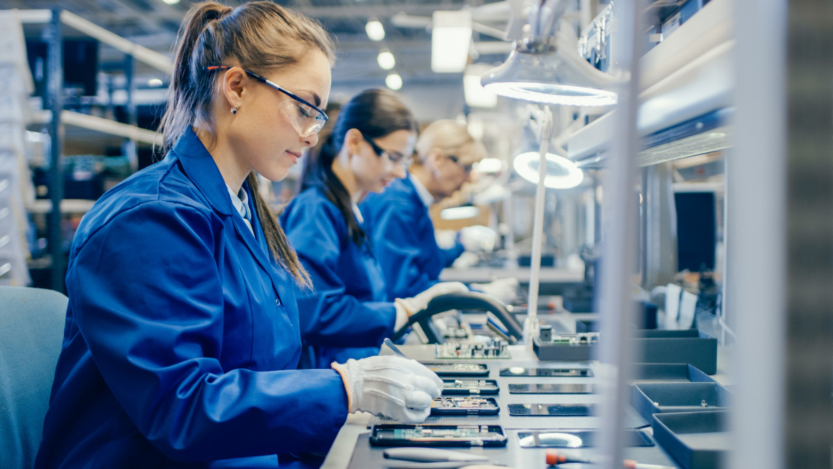 Female Electronics Factory Workers in Blue Work Coat and Protective Glasses Assembling Printed Circuit Boards for Smartphones with Tweezers. High Tech Factory with more Employees in the Background.