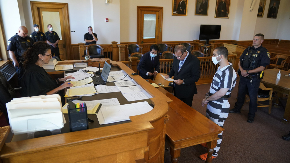 Hadi Matar 24 éves New Jersey-i férfi, Salman Rushdie indiai származású brit-amerikai író merénylője (j2) kirendelt ügyvédje, Nathaniel Barone (k) társaságában vád alá helyezési eljárásán a Chautauqua megyei bíróságon, a New York állambeli Mayville-ben 2022. augusztus 13-án. Matart azzal vádolják, hogy késsel megtámadta Rushdie-t, mielőtt megkezdte előadását a Chautauquai Intézetben augusztus 12-én. Rushdie-ra 1989-ben Irán akkori vallási vezetője, Ruhollah Khomeini ajatollah iszlám halálos ítéletet, fatvát mondott ki az egy évvel korábban megjelent, Iránban istenkáromlás miatt betiltott könyve, a Sátáni versek miatt.
