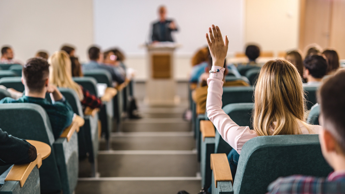 Back view of female college student raising her hand to answer the question on a class at lecture hall.