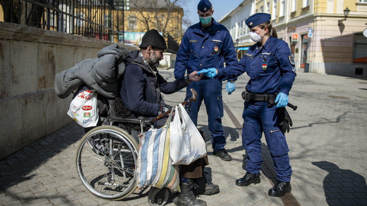 Rendőrök gumikesztyűt adnak egy kerekesszékes férfinak az egri Széchenyi utcában a kijárási korlátozás kezdetének napján, 2020. március 28-án. A koronavírus-járvány miatt kijárási korlátozást vezettek be Magyarország egész területére március 28. és április 11. között. Ebben a két hétben az otthonokat, a lakóhelyeket csak munkavégzés vagy az alapvető szükségletek ellátása céljából lehet elhagyni. Az élelmiszerboltokat, drogériákat, patikákat 9 és 12 óra között kizárólag a 65 év felettiek látogathatják. A rendőrség a közterületeken megnyugtató, segítő, empatikus és lojális intézkedésekkel sarkallja a lakosságot a kijárási korlátozás betartásra.
