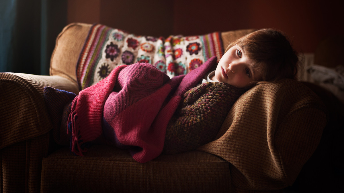 This is a picture of a 7 year old girl lounging on an armchair. She has blue eyes and light brown hair. She is looking into the distance with a wistful expression on her face. She has a blanket over her. The room is dark around her.