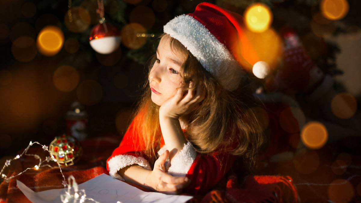 Little girl in a Santa hat and red dress under Christmas tree is dreaming, waiting for the holiday, lying on a plaid blanket. A letter on piece of paper, gifts. New year, Christmas. defocus lights