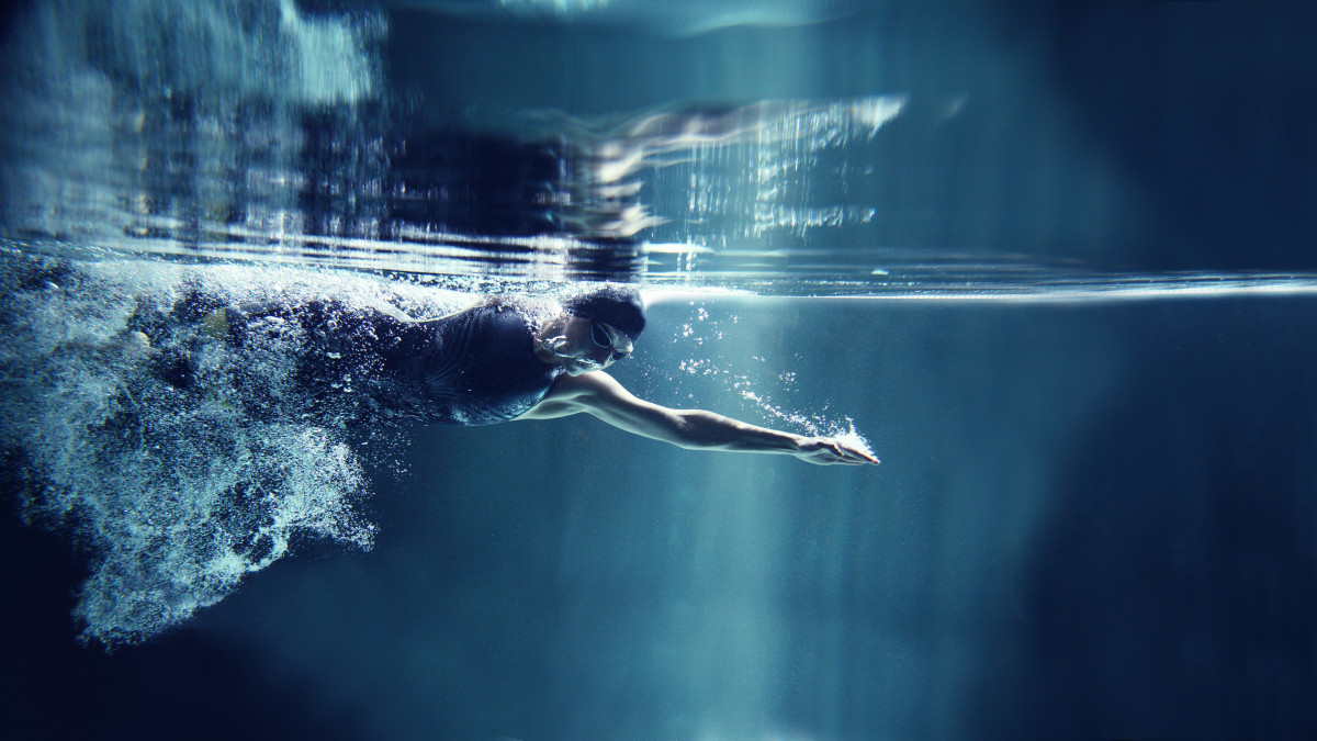 A female athlete is swimming crawl. She is wearing professional, black swim wear, swimming glasses and cap. You can see her torso, head and one hand. She is emerging from air bubbles. She is exhaling air to water and has one arm streatched in front. She is looking down. In the top of image you can see the surface of water and reflection of the scene. The background is dark blue. There are no swimming pool elements. This is a horizontal image.