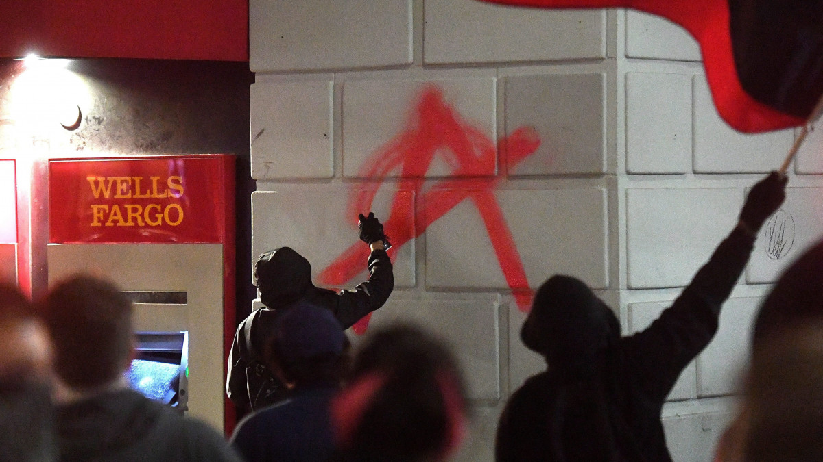 Berkeley, 2017. február 2.Egy tüntető az anarchizmus jelképét rajzolja festékszóróval a Wells Fargo bank egyik fiókjának falálra a kaliforniai Berkeley városban 2017. február 1-jén. A több száz tüntető követeltelésének engedve a Kaliforniai Egyetem (UC) berkeley-i részlegének vezetősége lemondta Milo Yiannopoulos, a Breitbart News jobboldali amerikai hírportál szerkesztőjének az egyetemen tartandó beszédét, miután a feldühödött tömeg számos környező épületben kárt tett. Yiannopoulos nyíltan támogatja Donald Trump amerikai elnököt. (MTI/EPA/Noah Berger)