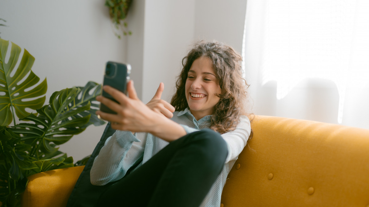Young caucasian woman smiling with thumbs up while taking a selfie or having a video call on her cell phone from her sofa at home. Technology concept.