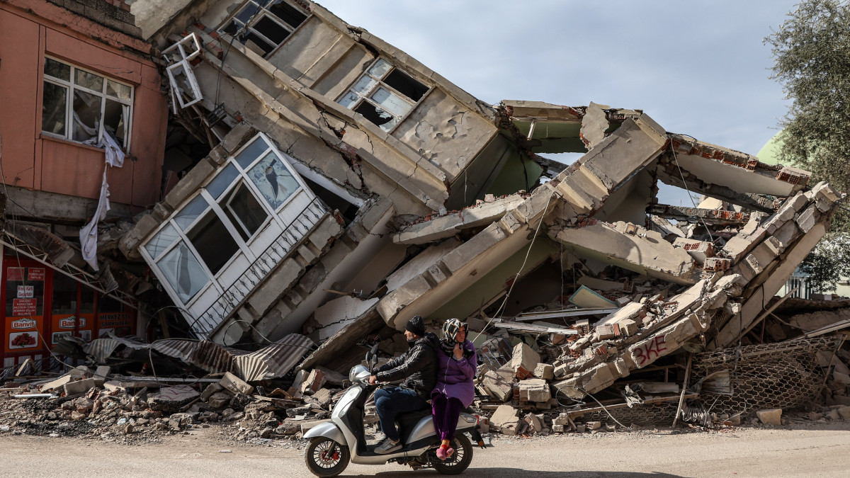Robogóval közlekedő helyi lakosok egy összedőlt épület romjai előtt a földrengés sújtotta délkelet-törökországi Adiyamanban 2023. február 19-én, tizenhárom nappal a Dél-Törökországot és Észak-Szíriát sújtó 7,7-es és 7,6-es erősségű két földrengés után. A természeti katasztrófa halálos áldozatainak száma meghaladja a 45 ezret.