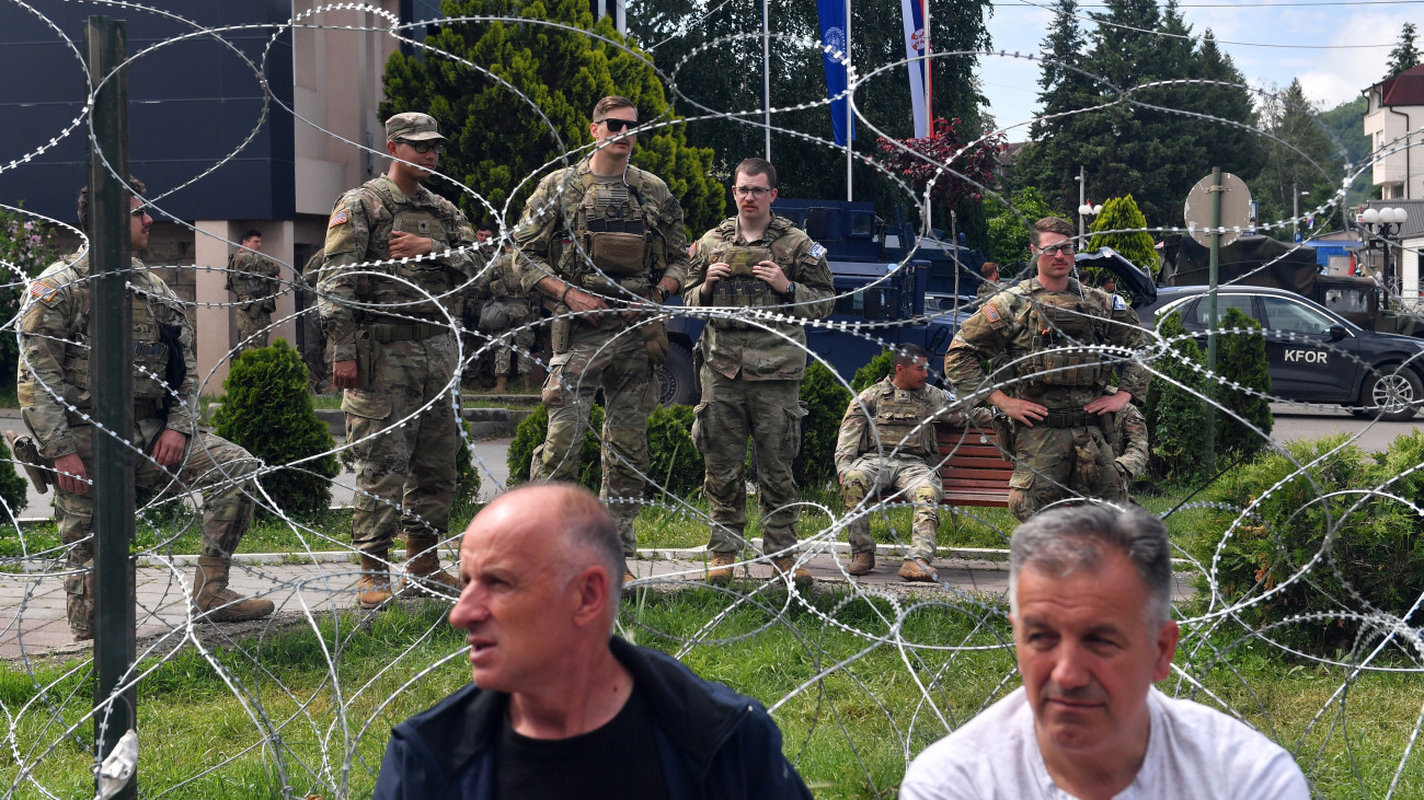 A NATO-parancsnokság alatt működő koszovói békefenntartó erő, a KFOR amerikai katonái a leposavici városháza előtt, az előtérben helybeli szerb férfiak 2023. június 2-án. Négy nappal korábban a KFOR 30 katonája, köztük 19 magyar, illetve 52 szerb tüntető megsebesült a többségében szerbek lakta másik észak-koszovói városban, Zvecanban kitört zavargásokban.