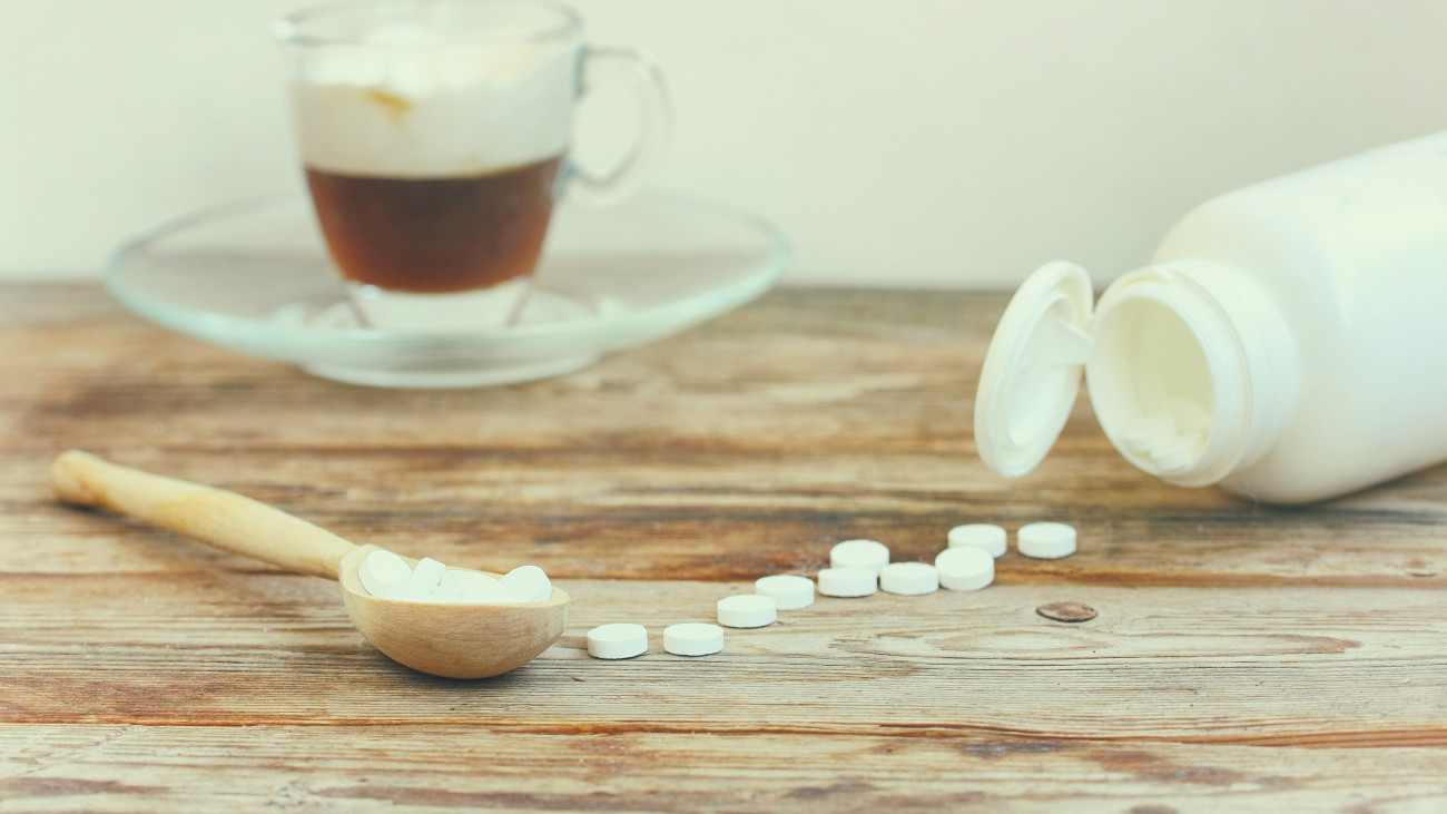 sweetener in tablets in wooden spoon on table, white jar with sweetener, cup of cappuccino coffee, selective focus