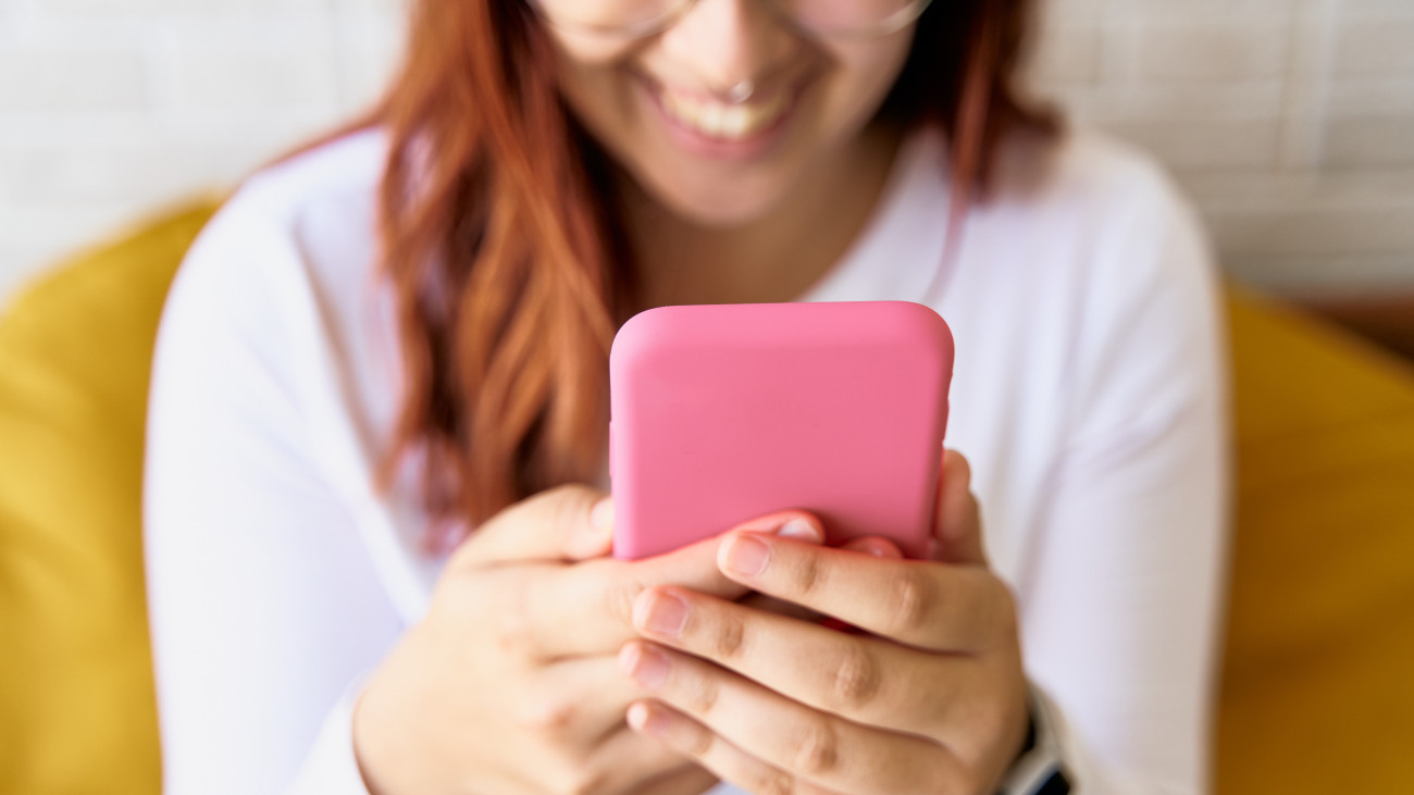 Irreconocible teenager texting with pink cell phone in white shirt and glasses with smile against yellow pillows