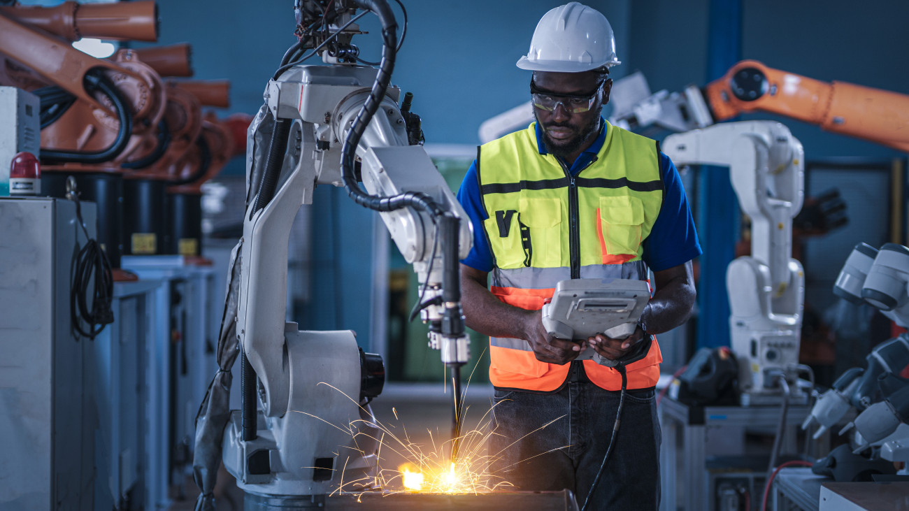African engineers control robotic arc welding at production line of factory, Male technician maintenance automatic robotic hand in smart factory, industrial and manufacturing concept