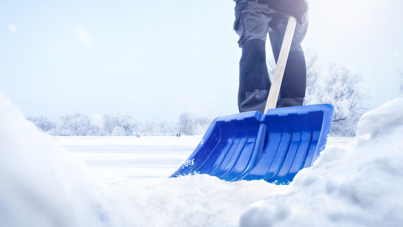 A person using a snow shovel in a snowy landscape. Only the lower part of the person is visible. Low angle shot with snow piles in the foreground.