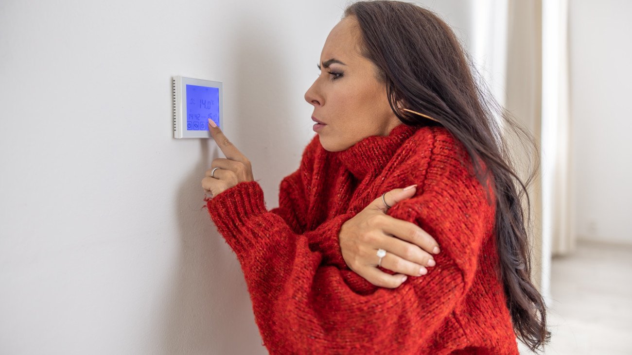 Freezing woman at home wears sweater and tries to raise the temperature on thermostat while energy crisis hits Europe in the winter.
