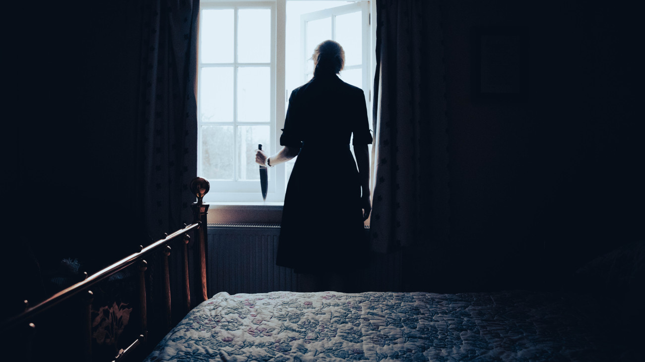 Lady with the knife standing in a bedroom looking out of the window with a knife, as if waiting for her victim