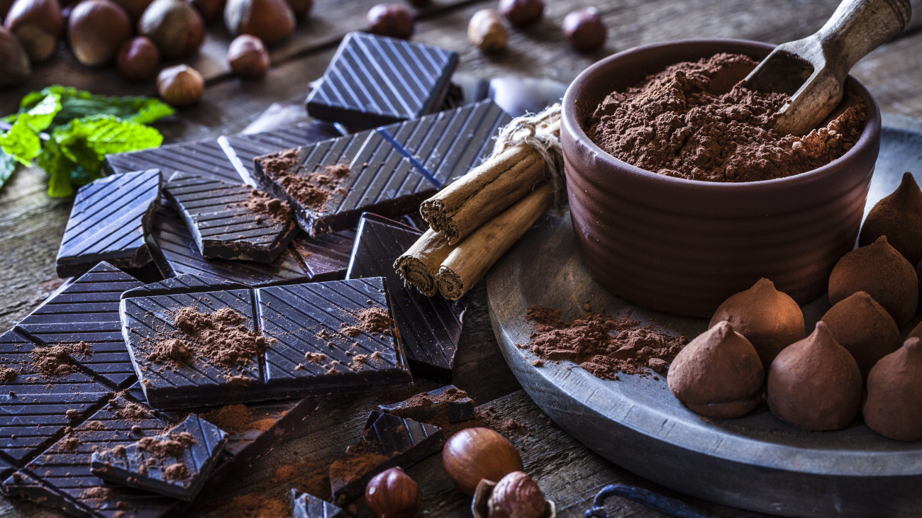 Rustic wooden table filled with ingredient for preparing homemade chocolate truffles. The composition includes dark chocolate truffles, chocolate bars, cocoa powder, cinnamon sticks, vanilla beans, mint leaves and hazelnuts. Predominant color is brown.