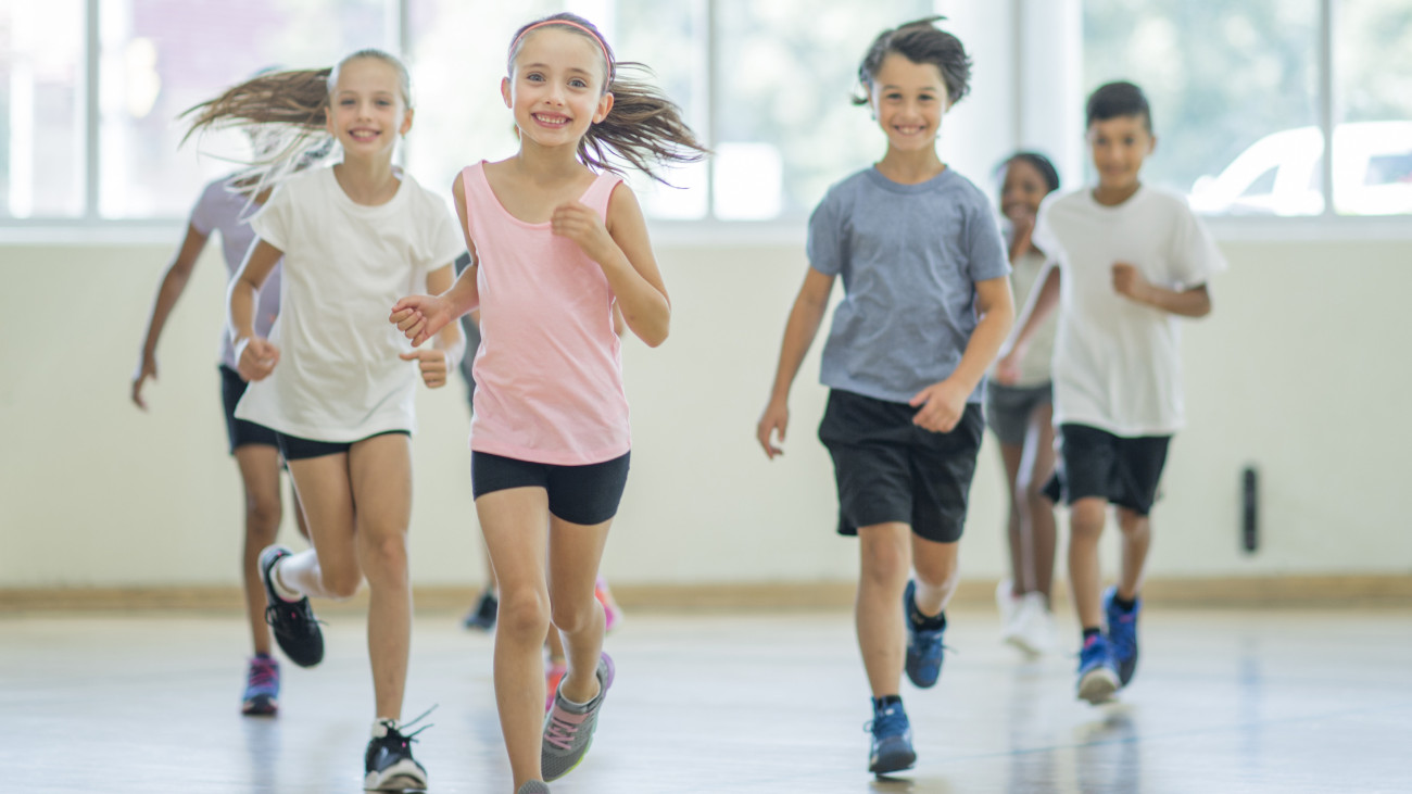 A multi-ethnic group of children are indoors in a health center. They are wearing casual clothing and running shoes. They are running towards the camera.