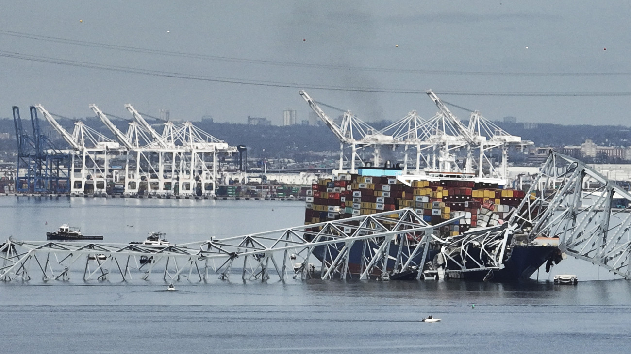 Konténerszállító hajó a baltimore-i Francis Scott Key híd acélszerkezetének roncsai alatt 2024. március 26-án, miután az éjjel a teherhajó az acél tartóoszlopnak ütközött, és a híd összeomlott. Eddig két embert mentettek ki a Patapsco folyóból, hat másikat még keresnek. Víz alatti radarok több járművet jeleztek a folyó mélyén.